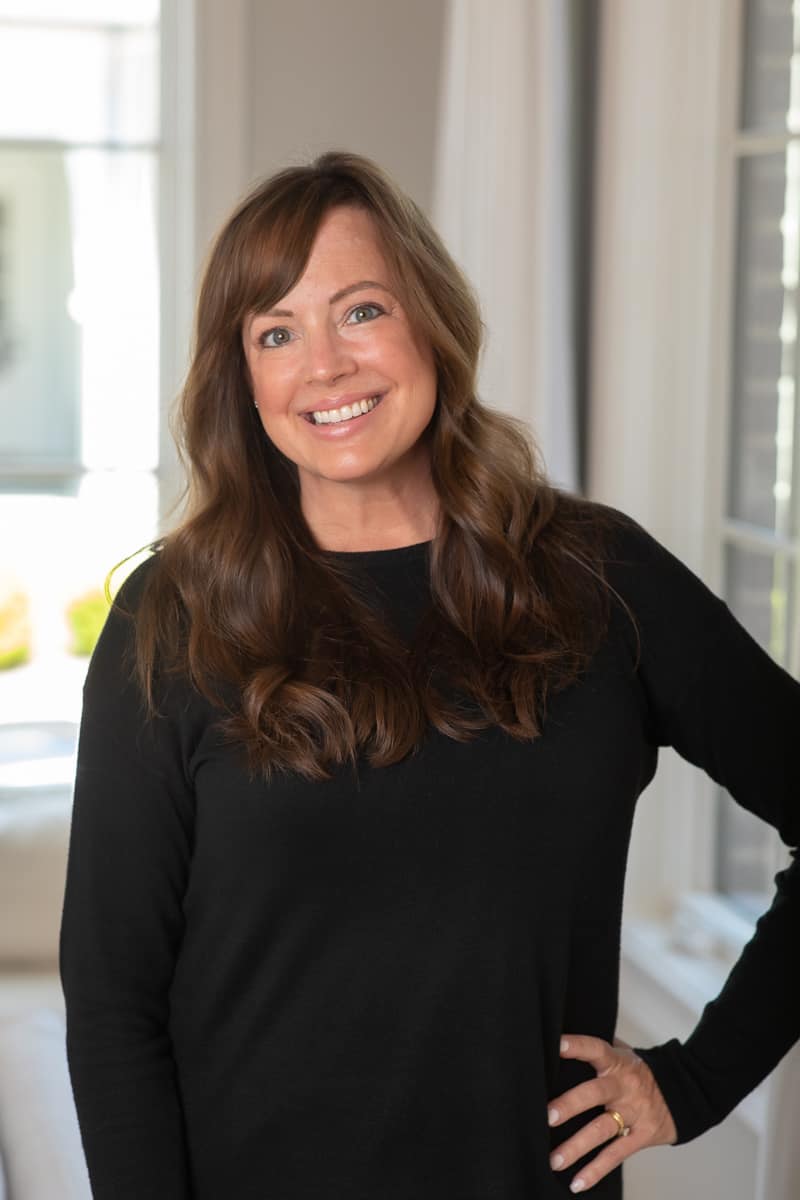 Woman in black sweater top poses with hand on hip in residential area with window behind.